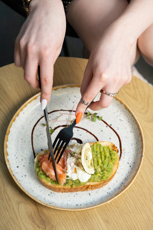 Toast with salmon and a poached egg on an avocado spread. Female hands with a fork.
