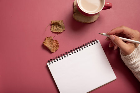 Table desk with notepad and female hand wit pencil and fall leaves on burgundy colored background. Autumn flat lay composition.の写真素材