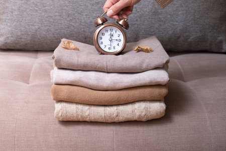 Stack of cozy knitted sweaters with autumn leaves and alarm clock in women's hands on the background of the sofa. Warm clothes time concept.の写真素材