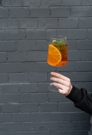 Close-up of woman's hand holding glass of cocktail with orange slice on black brick wall background.の写真素材