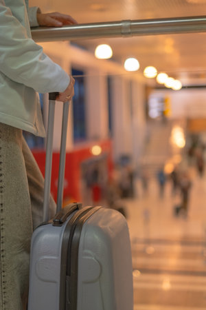 A traveler stands by a rail holding a suitcase, observing the busy airport terminal. Multiple passengers are seen walking past, creating a lively atmosphere during peak travel hours.の写真素材