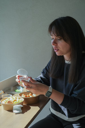 In a well-lit cafe, a woman sits at a table focused on a takeout container while enjoying her meal....