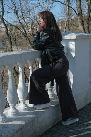 A young woman leans elegantly against a white railing, enjoying a sunny day in a park. She wears a black leather jacket and stylish pants, surrounded by trees.の写真素材