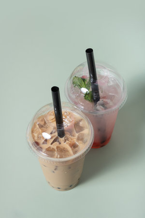Two bubble tea drinks sit side by side, one pink with mint and the other creamy brown, both filled with ice and topped with black straws, perfect for a refreshing treat.の写真素材