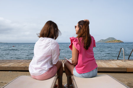 Two friends sit on lounge chairs, facing the ocean, enjoying a peaceful day by the sea. Soft waves lap against the shore, creating a serene atmosphere under a clear sky.の写真素材
