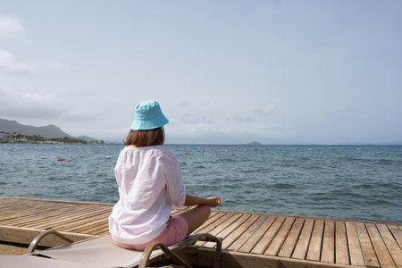 A person enjoys a tranquil moment by the water, seated on a wooden dock. The calm sea stretches out towards distant mountains, with gentle waves lapping at the shore under a clear sky.の写真素材