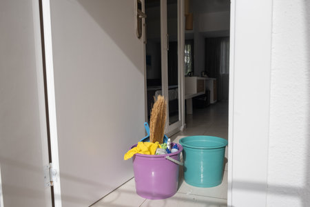 Cleaning tools, including a broom and colorful buckets, rest outside a doorway leading to an interior space. The bright sunlight suggests a warm day by the sea.の写真素材