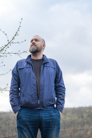 Man standing confidently outdoors in a blue jacket under a cloudy sky near a scenic landscape with rolling hillsの写真素材