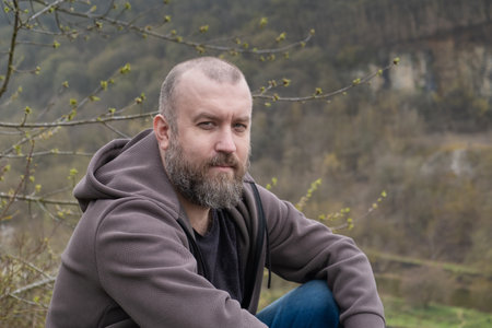Man 40 year old with beard sitting on hillside in nature with trees and mountains in the backgroundの写真素材
