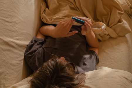 A teenage boy is lying on a cozy bed, focused on his smartphone while wrapped in blankets. The warm afternoon light fills the room, creating a relaxed atmosphere.の写真素材