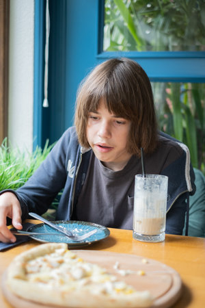A 13 year old boy sits at a cafe table, focused on his dessert and drink. Surrounded by green plants, he appears to savor the moment in a cozy afternoon setting.の写真素材