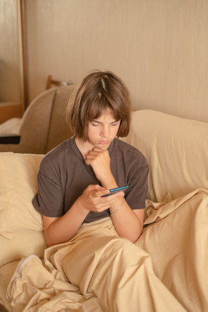 A boy around 13 years old is seated comfortably on a sofa, focused on his smartphone while wearing a relaxed outfit. The light-colored surroundings create a calm atmosphere.の写真素材