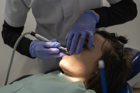 A dental professional is cleaning a young patient's teeth in a bright, modern clinic. The patient is seated and relaxed in a chair, wearing a bib.の写真素材