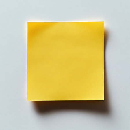 A bright yellow sticky note lies on a smooth white surface, ready for reminders.の素材