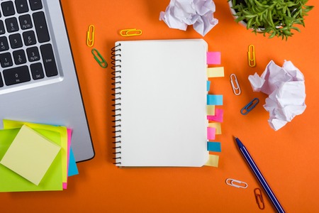 Office table desk with set of colorful supplies, white blank note pad, cup, pen, pc, crumpled paper, flower on orange background. Top view and copy space for text.の写真素材