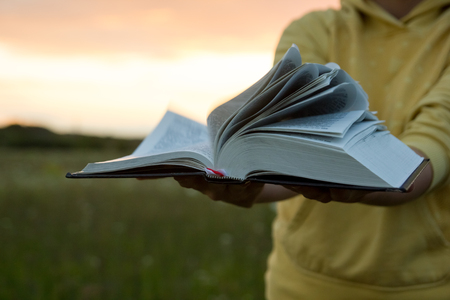 Female hands holding opened hardback book, diary with fanned pages on blurred nature landscape background against sunset sky with back light. Copy space, back to school education concept.の写真素材
