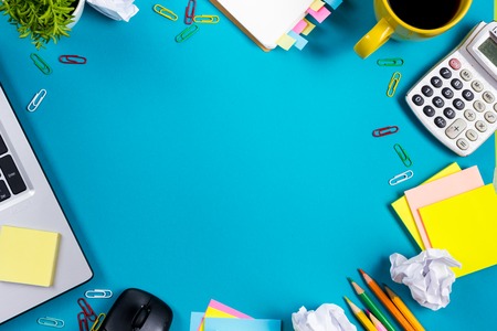 Office table desk with set of colorful supplies, white blank note pad, cup, pen, pc, crumpled paper, flower on blue background. Top view and copy space for text.の写真素材