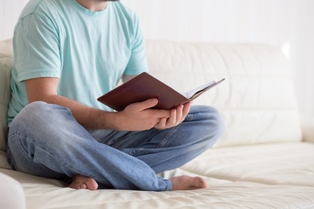 Man reading old hardback note book holding in his hands, on the white couch. Domestic lifestyle. Education concept.の写真素材
