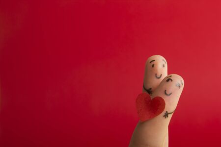 Valentines day fall in love concept. Painted happy funny fingers smiley holding red heart against red background with copy space for ad textの写真素材