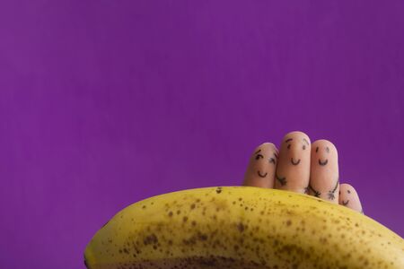 Painted funny fingers smiley holding yellow banana against purple backgroundの写真素材
