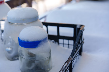 this unique photograph shows glass jars placed in a black plastic box in a snowy environment. The jars are partially covered in snow, creating a magical and mysterious winter scene. This image perfectly highlights the beauty of winter and the contrast between the cold snow and the warmth stored in the jars.の写真素材