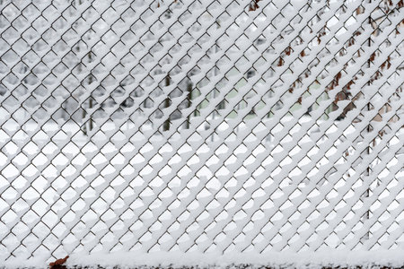 A chain link fence covered in snow photographed from the front, snow stuck to and frozen into the fence details, a yard photographed through the fence during a snowfall, weather conditions snowfall, from a close distanceの写真素材