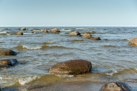 rocky stretch of coast, Baltic Sea large stones are scattered in the water and along the shore, creating a rough but natural landscape. In the foreground, bright green water grass grows between the stones, the water is wavy and reflects the gray-blue skyの写真素材