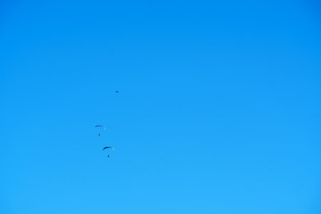 A wide shot of a vast, clear blue sky featuring three individuals engaged in paragliding. The small silhouettes of the paragliders with their colorful canopies are visible against the vibrant backdrop, suggesting freedom, adventure, and outdoor sports. The expansive sky offers ample copy spaceの写真素材