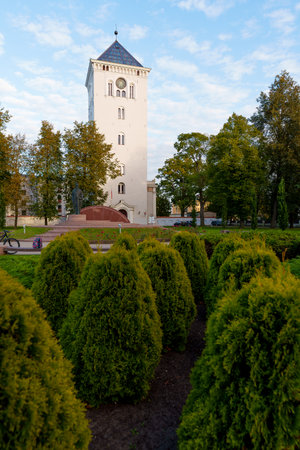 The white tower of Jelgava's landmark church with a clock, framed by vibrant green park shrubs and trees under a blue sky, representing history and urban greeneryの写真素材