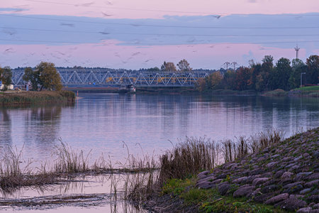 A serene landscape featuring a steel railway truss bridge spanning a calm river under a soft,...