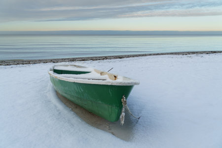 A quiet winter scene featuring an abandoned  fishing boat resting on a snow-covered sandy beach with a small bare tree, framed by a calm sea and forest horizonの写真素材