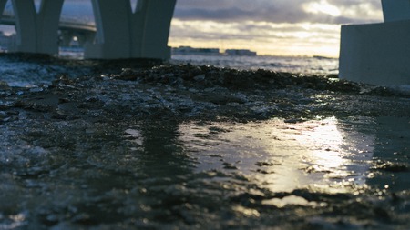 Close up picture of beach's surface. Ice is melted. Some concrete constructions are on a background. Sky is covered by clouds. Some sun lights reflects on melted surface of ice cover. Waves comes.の写真素材