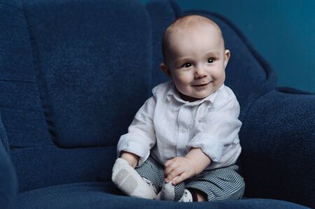 Toddler in the white shirt sitting on the blue sofa and touching his feets with hands