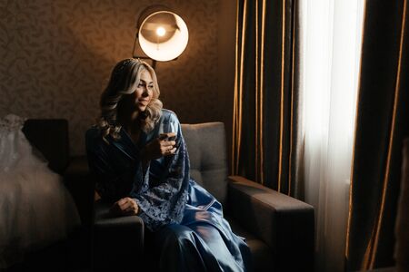 Blonde woman in the blue peignoir with the glass of champagne in her hands. Bride portraitの写真素材