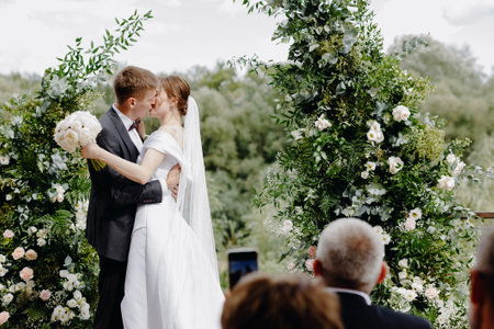 Happy newlyweds stand at the wedding ceremony near the wedding arch and kiss. High quality photoの写真素材