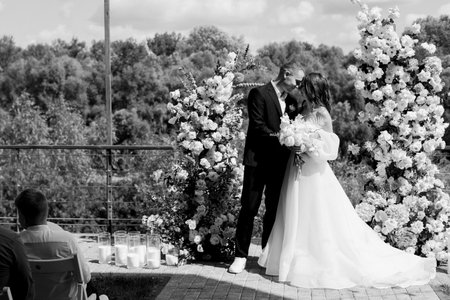 A stylish groom and a beautiful bride in a long dress are standing near an arch of fresh flowers. Black and white photo. High quality photoの写真素材