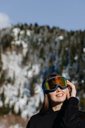 A girl in ski goggles and a black suit poses against the backdrop of snow-capped mountains.の写真素材