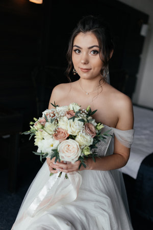 The bride sits on the bed in a dress, holding a bouquet and looking at the cameraの写真素材