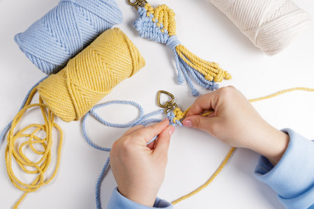 Hands of young woman working on macrame decor with blue and yellow threads over creative work table,...