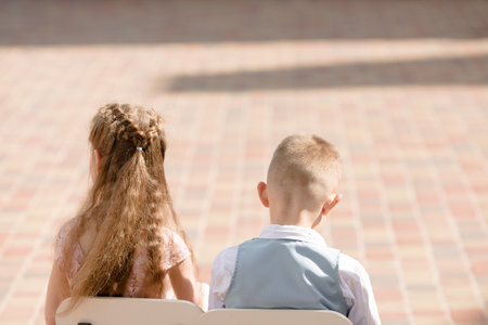 Back portrait of a boy and a girl of 6 years old sitting on chairs outside on a tile backgroundの写真素材