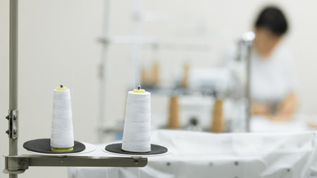 Two skeins of white sewing threads and a large spool placed on a table with a blurred working woman...