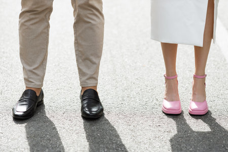 A trendy couple poses on a sunny pavement, showing off their fashionable footwear and outfits. The woman wears a white dress and pink heels, while the man wears beige trousers and black shoesの写真素材