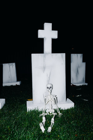 A skeleton sits in front of a tombstone cross at night, creating a spooky atmosphere.の写真素材