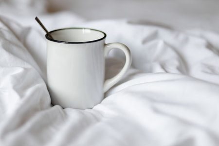 A white mug with a spoon, resting on a soft, white bed, evoking a sense of warmth and comfort.の写真素材