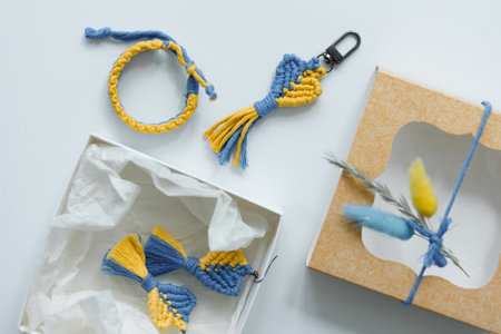 A flat lay of handcrafted macrame accessories in blue and yellow, including a bracelet, keychain, and earrings displayed beside an elegantly wrapped gift boxの写真素材