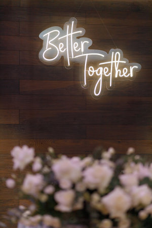 A captivating image capturing the warm, inviting glow of a Better Together neon sign affixed to a wooden wall. Below, blurred flowers bloom, adding a touch of natural beauty to the modern aestheticの写真素材