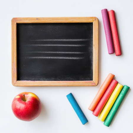 A vibrant arrangement featuring a mini chalkboard surrounded by colorful chalk sticks and a shiny red apple on a pristine white surface. This setup invites creativity and exploration.の素材