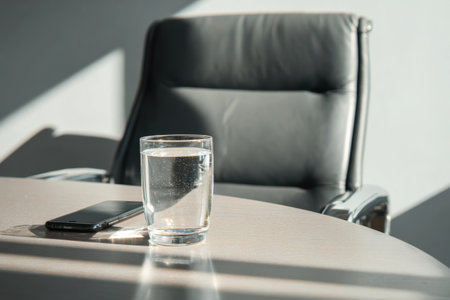 A minimalist workspace features a glass of refreshing water and a smartphone resting on a side table. Sunlight casts gentle shadows, creating a calm and focused atmosphere for productivity.の素材