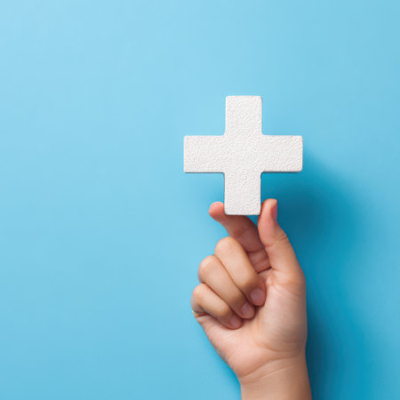 A hand gently holds a white medical cross symbol, set against a vibrant blue backdrop, symbolizing health and care.の素材