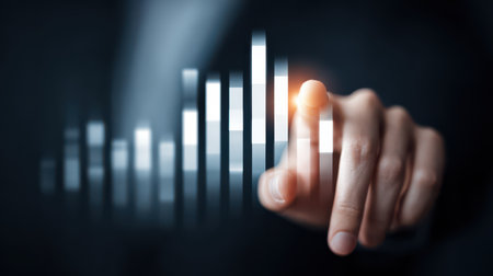 A hand gently touches a glowing bar on a data chart, symbolizing growth and progress in analytics, against a blurred dark background.の素材